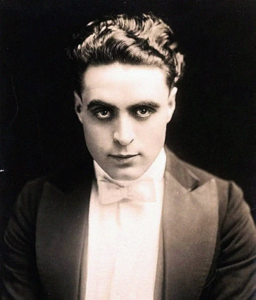 A young man with wavy dark hair, wearing a tuxedo and bow tie, stares intently at the camera against a dark background in a vintage black-and-white portrait.