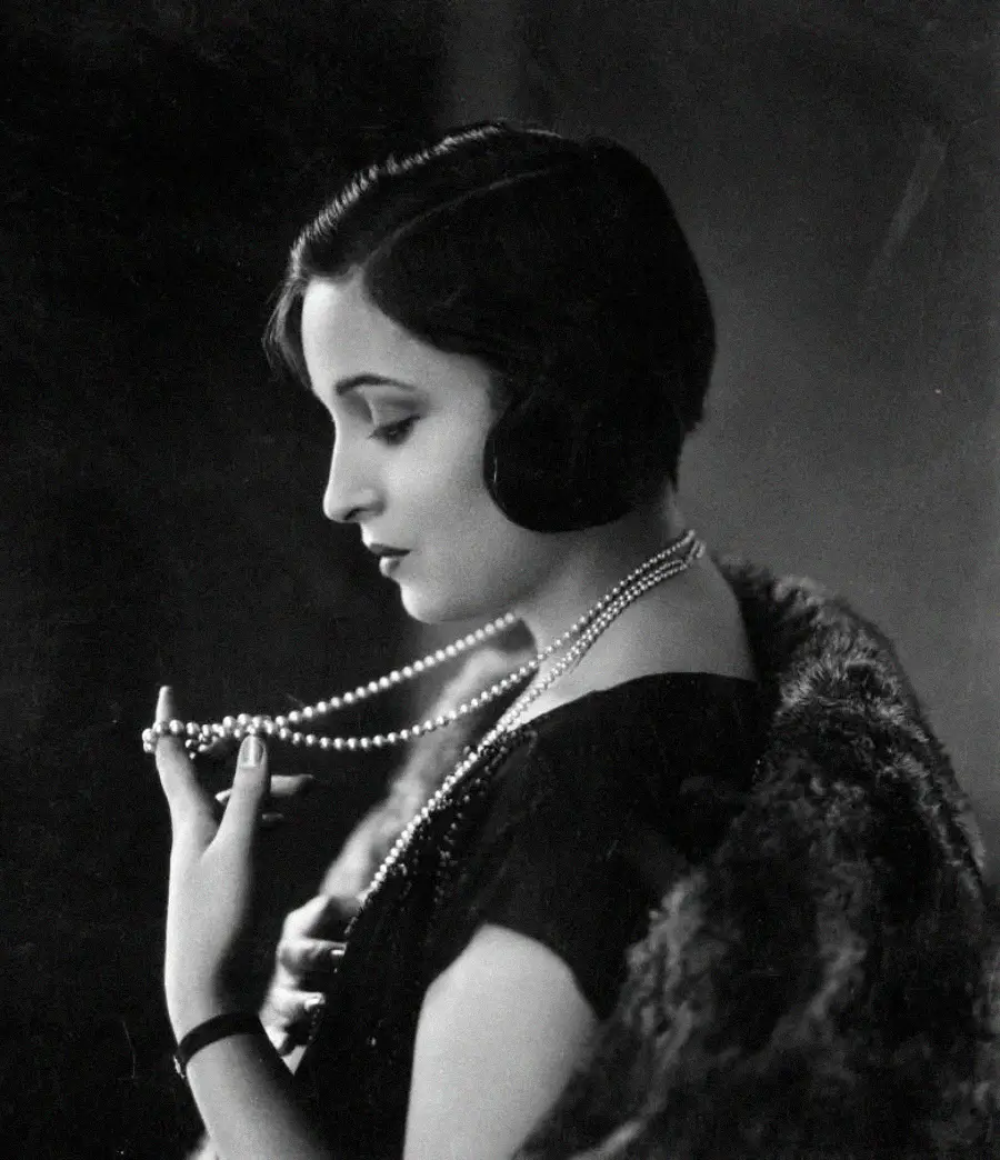 A woman in vintage attire poses in profile, wearing a fur stole and holding a long strand of pearls. She has short, dark, wavy hair and is set against a dark, softly lit background.