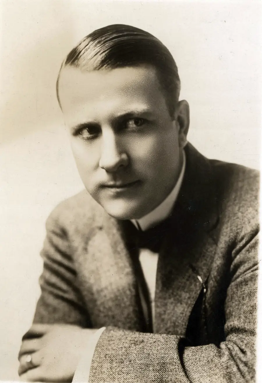 A black and white portrait of a man with neatly combed hair, wearing a suit, bow tie, and dress shirt, looking slightly to the side with a serious expression. His arms are resting in front of him.