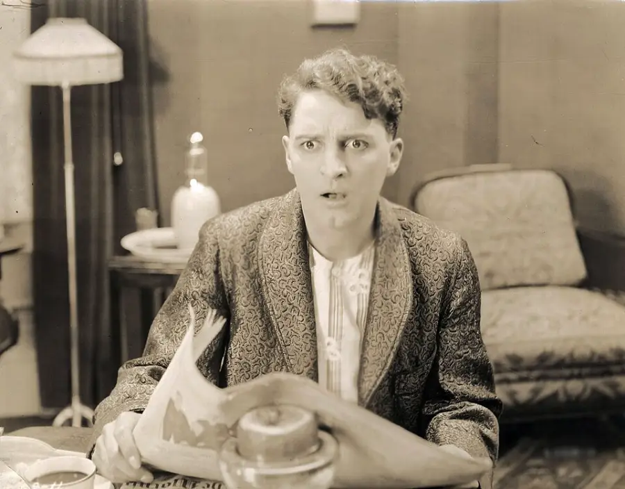 A man wearing a patterned jacket sits at a table, looking surprised or shocked while holding a newspaper. The room features vintage furniture, including a lamp, armchair, and a teapot on the table.