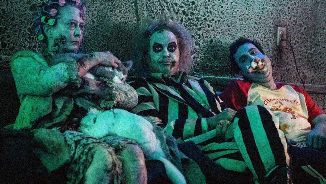 Three people in Halloween costumes sit on a couch: one with curlers and zombie makeup holding a cat, one dressed as Beetlejuice with wild hair and striped suit, and one in clown makeup with a toy over their nose.