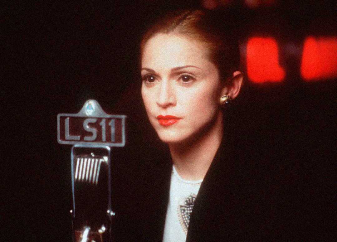A woman with light skin, wearing a black coat and red lipstick, sits in front of an old-fashioned microphone labeled "LS11." The background is dimly lit with red lights visible.