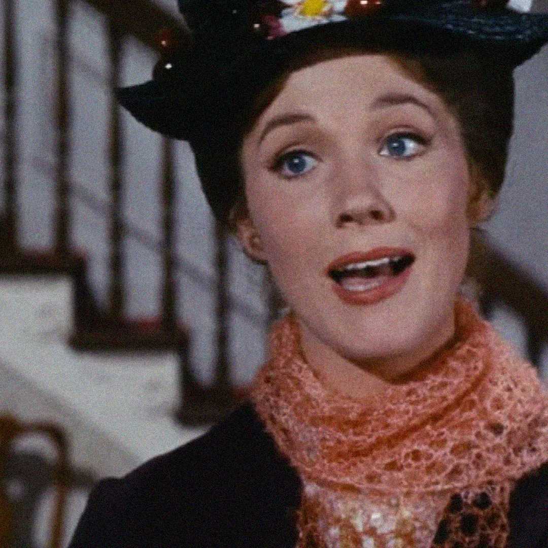 A woman wearing a dark hat decorated with flowers and a pink lace scarf smiles while standing in front of a staircase.