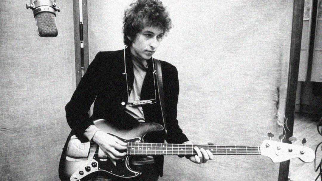 A black and white photo of a person with curly hair playing an electric bass guitar in a recording studio, wearing a harmonica holder around their neck and standing near a large microphone.