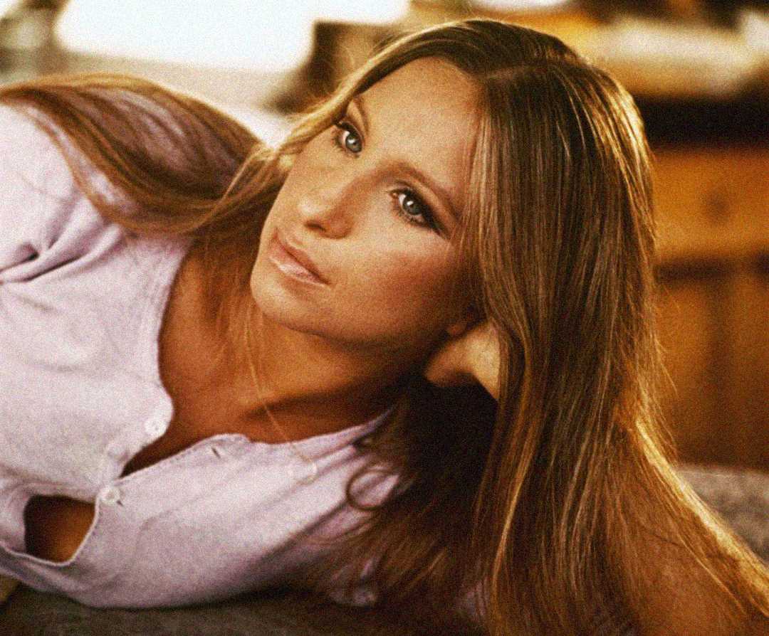 A woman with long, straight brown hair and light eyes lies on her side, resting her head on her hand and gazing thoughtfully into the distance, wearing a light lavender top. The background is softly blurred.