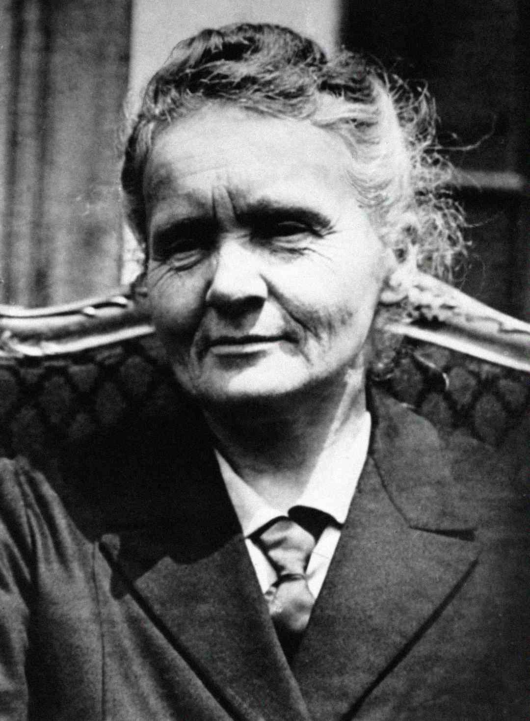 An older woman with light hair pulled back, wearing a dark jacket and blouse, sits on a patterned chair, looking slightly to the side with a calm expression.