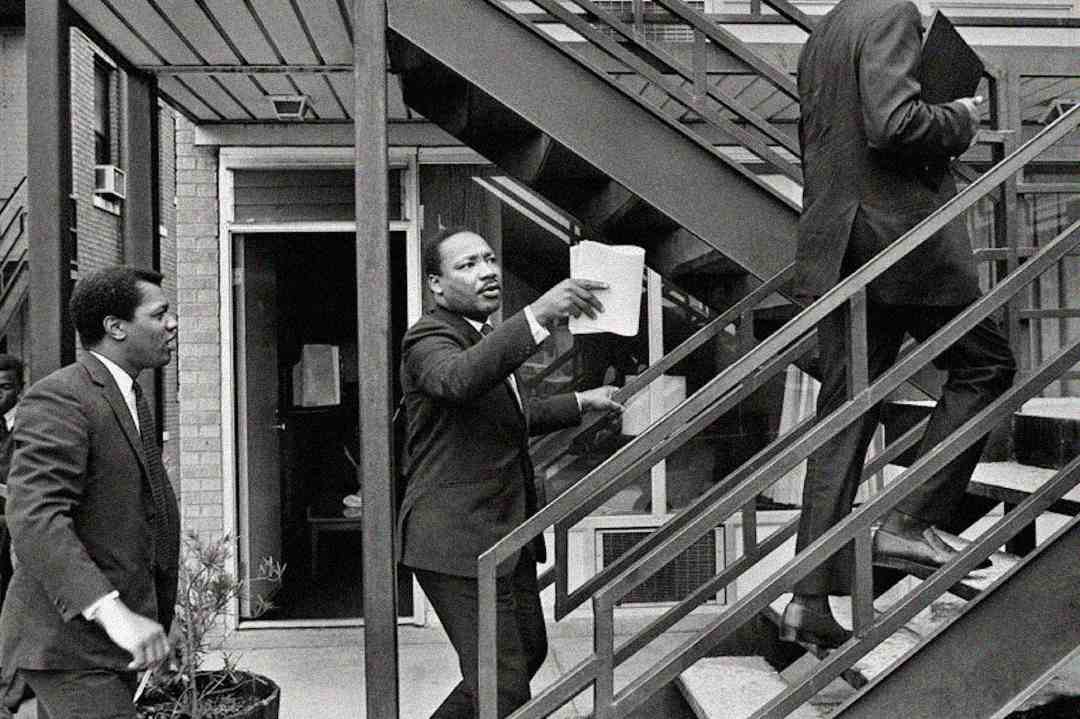 Three men in suits walk up an outdoor staircase attached to a brick motel. One man in the center, holding papers, looks upward while others follow behind him. It is a black and white photo.