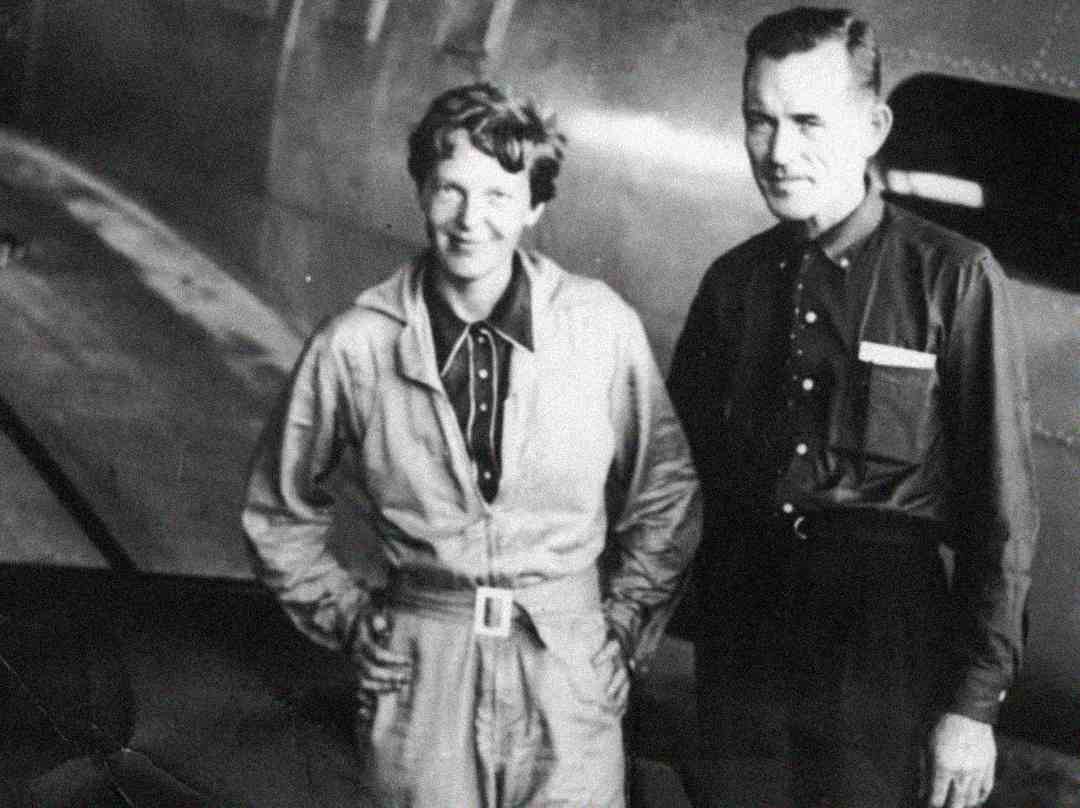 A woman in a flight suit stands beside a man in a button-up shirt and trousers, both smiling, in front of an aircraft. The image is black and white and appears to be historical.
