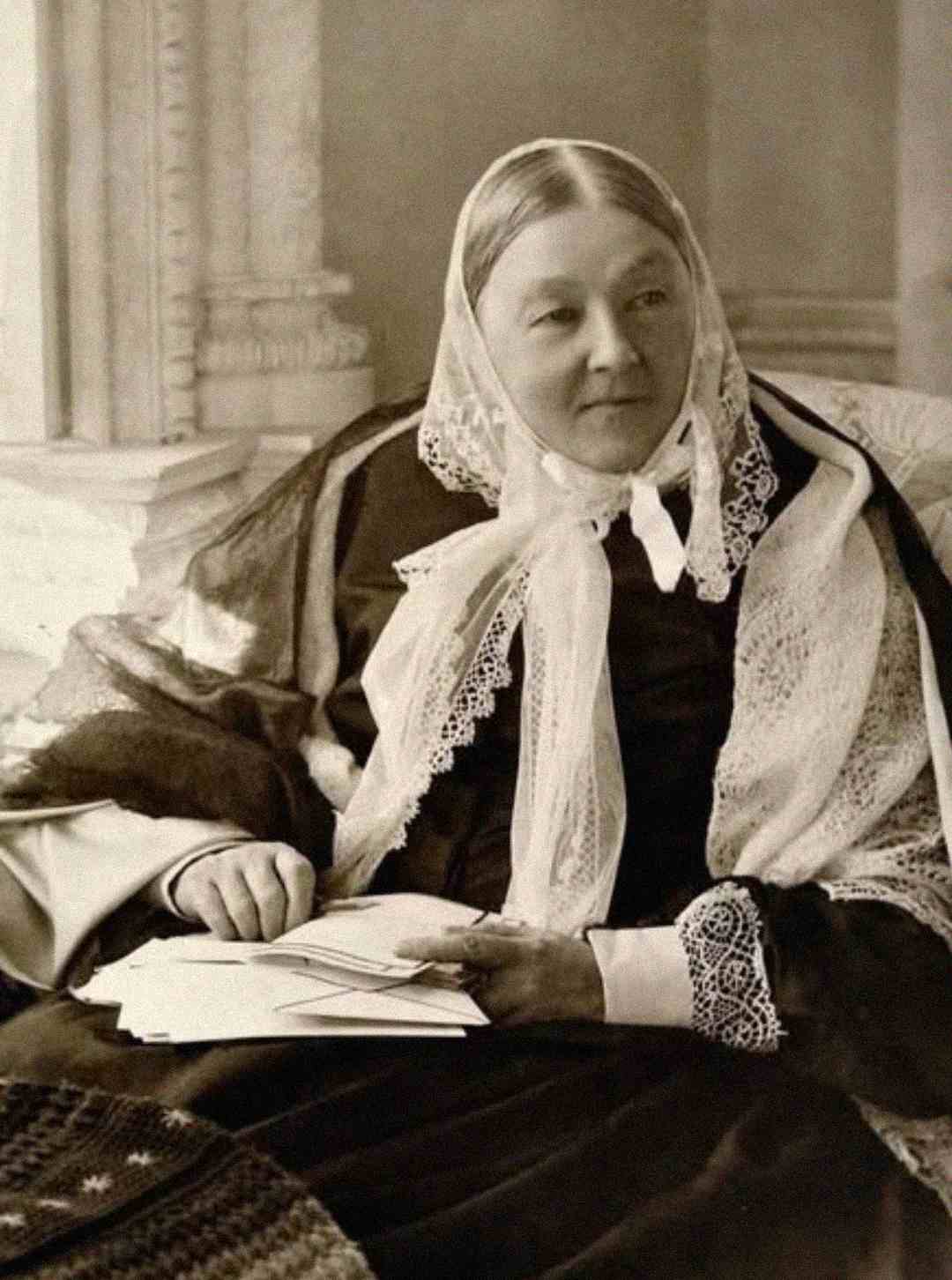 An older woman dressed in Victorian-era clothing and a lace headscarf sits, holding papers in her hand, with an ornate background behind her.