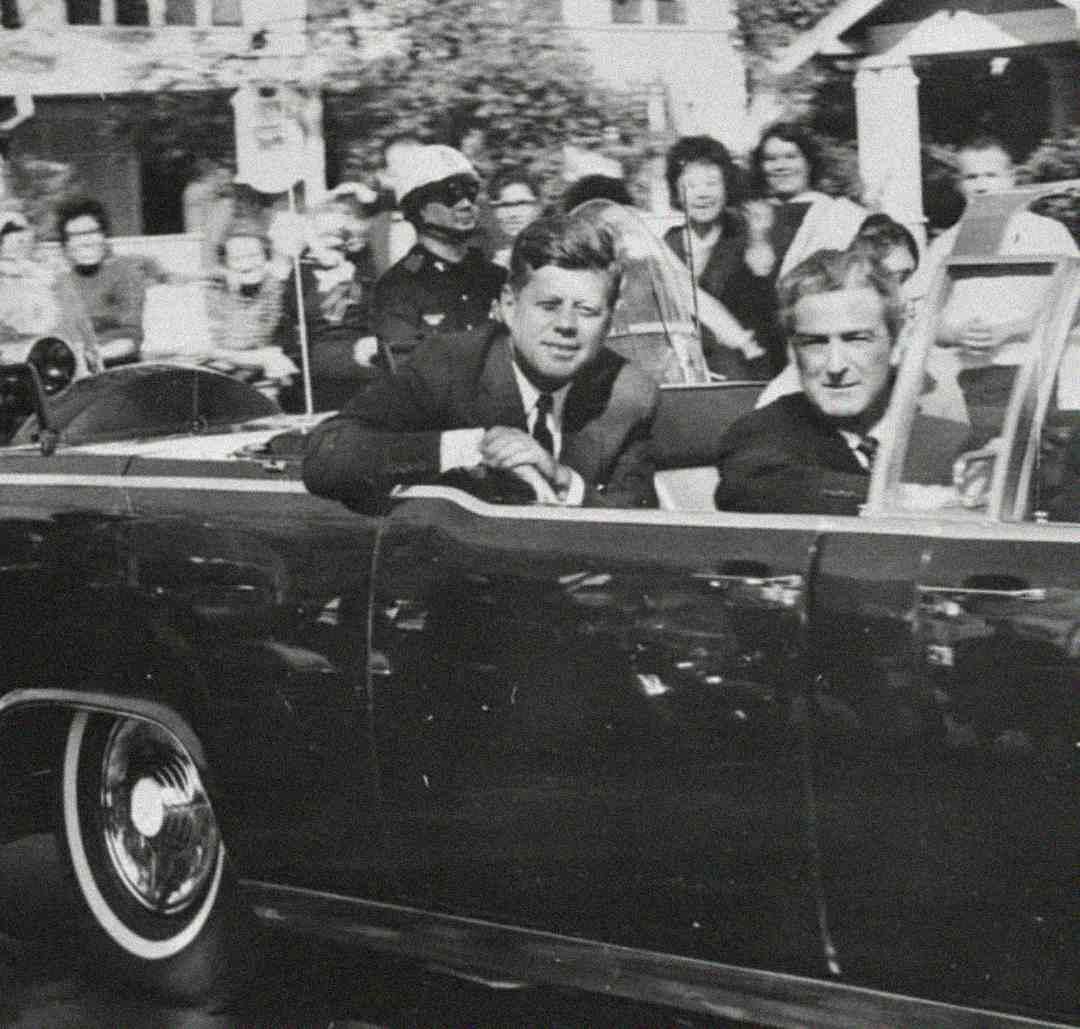 A black-and-white photo of two men in suits riding in a convertible car during a parade. Crowds of people watch from the sidewalk, some smiling and waving. One man looks directly at the camera, while the other looks ahead.