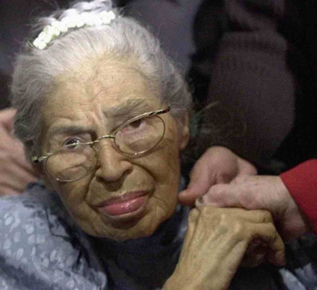 An elderly woman with gray hair, glasses, and a white hair clip is wearing a blue patterned shawl. She is holding someone's hand and looking slightly to the side with a calm expression.