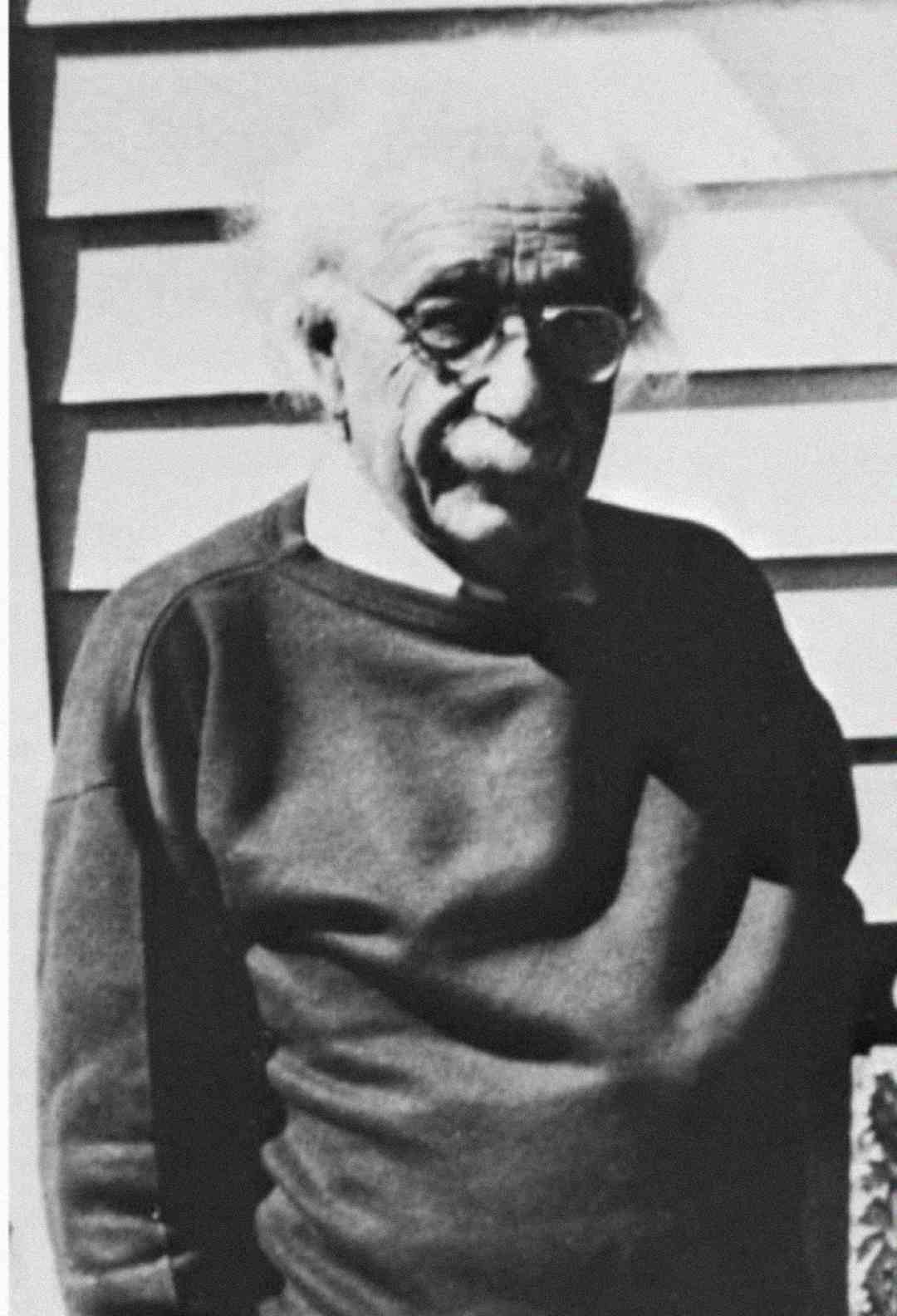 Black-and-white photo of an elderly man with wild white hair, glasses, and a mustache, wearing a dark sweater and standing in front of a house with siding.