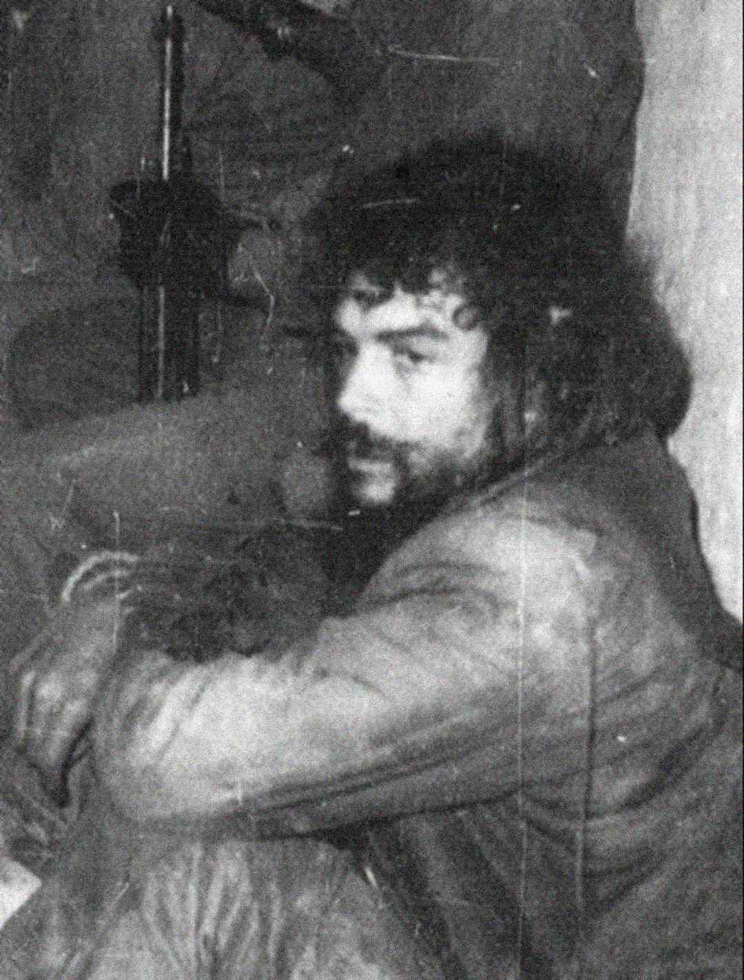 Black and white photo of a man with curly hair and a beard, sitting with his arms resting on his knees, looking at the camera. The image appears old and grainy, and a rifle is faintly visible in the background.