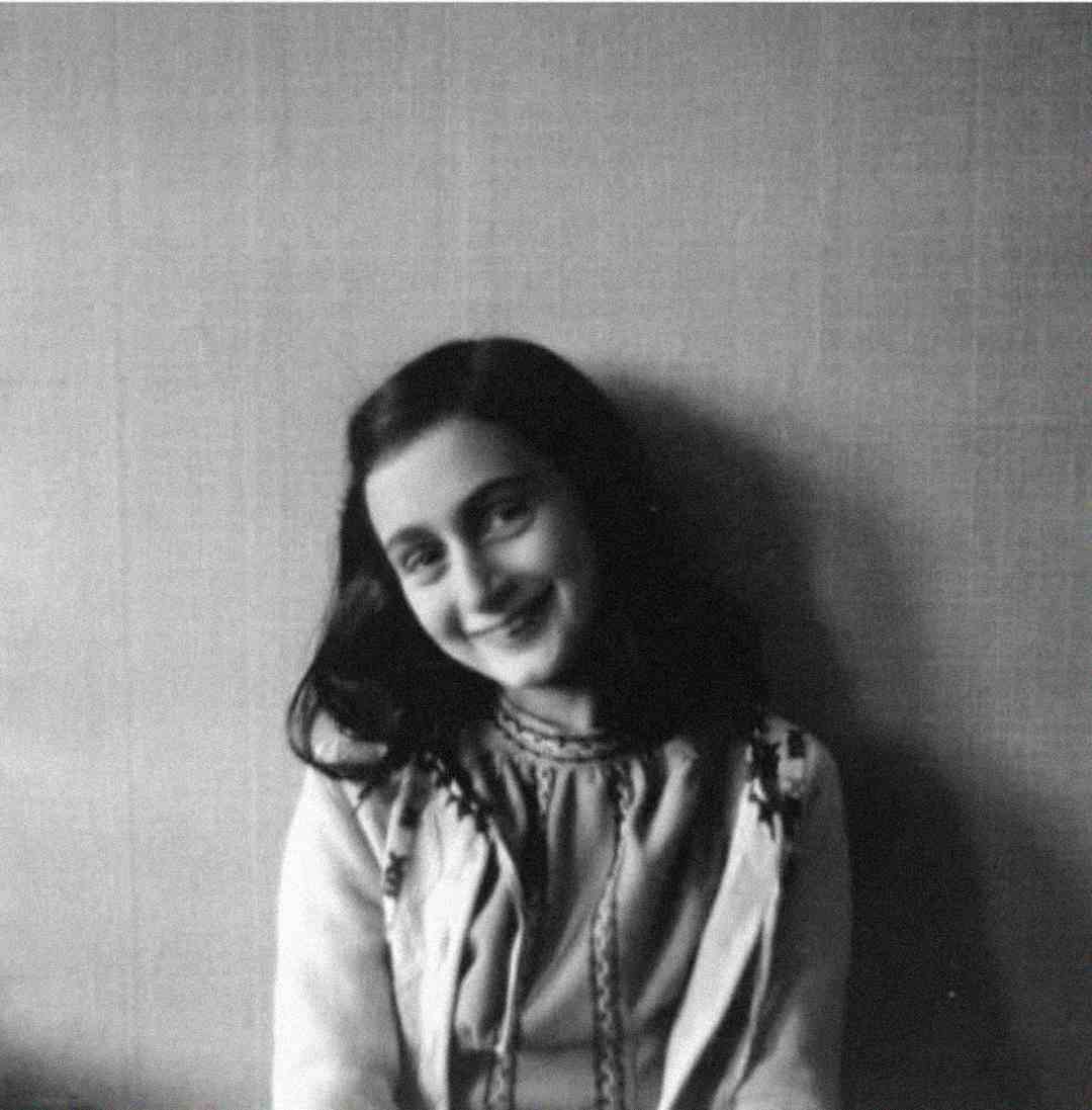 A black-and-white photo of a young girl with dark, wavy hair, wearing a light-colored blouse and smiling gently while sitting against a plain wall.