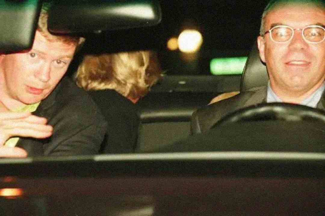 Two men sit in the front seats of a car at night, one on the left gesturing with his hand and the other on the right wearing glasses. A person with blonde hair sits in the back seat, facing away.