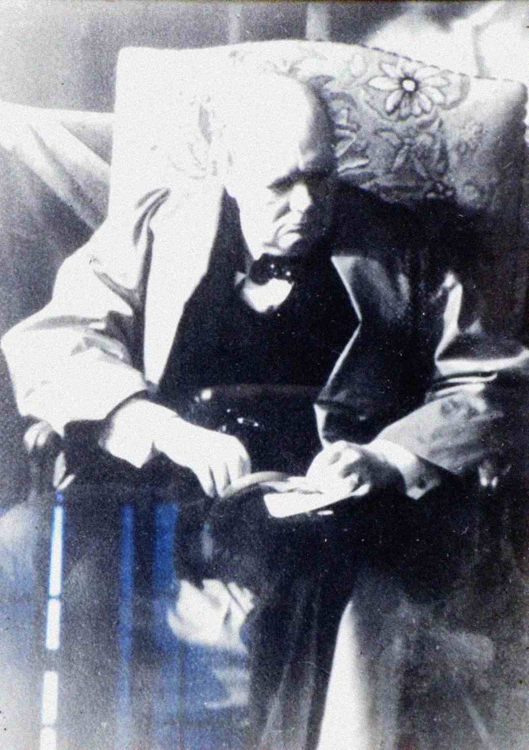 An elderly man in a suit sits in an armchair, looking down and reading a book. The image is black and white and has a soft, blurry quality. A patterned cushion is behind him.