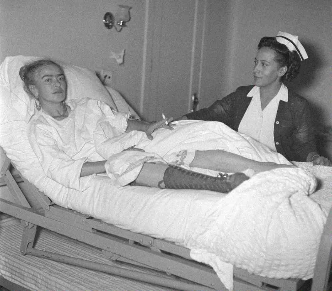A woman lies in a hospital bed with her leg in a cast while a nurse sits beside her, holding her arm, in a hospital room setting.