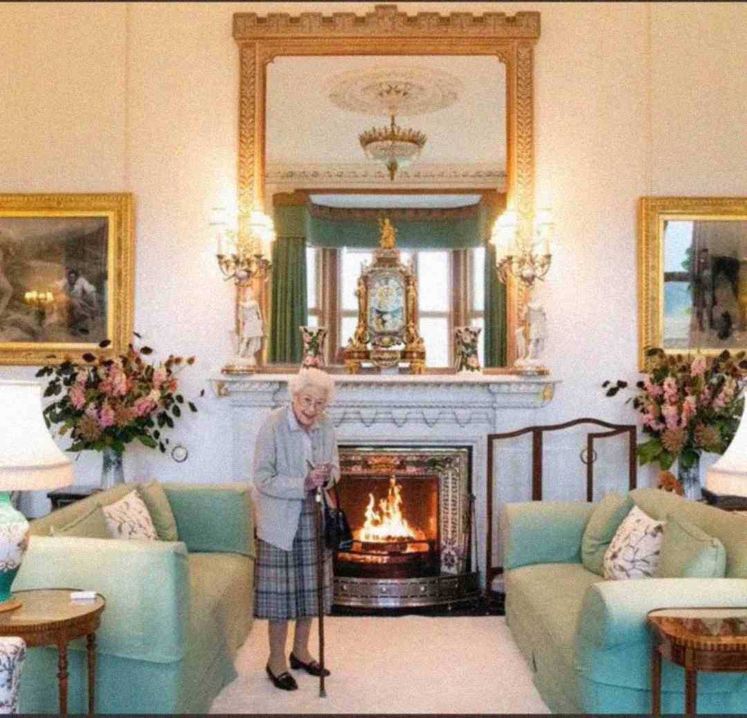 An elderly woman with a cane stands in an elegant living room with pastel sofas, a lit fireplace, large gold-framed mirror, ornate clock on the mantel, chandeliers, and floral arrangements.