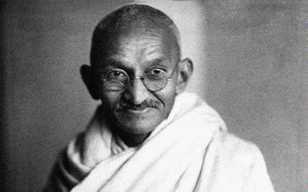 An elderly person with round glasses, a bald head, and a mustache wears a simple, draped white cloth and smiles gently while looking at the camera. The background is plain and out of focus.