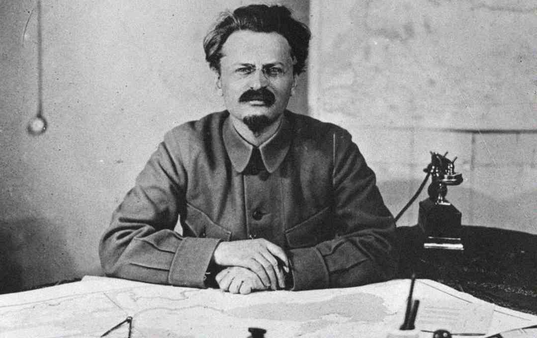 A man with glasses, a goatee, and dark hair sits at a desk covered with papers and a map, looking directly at the camera. There is a vintage telephone and a map on the wall behind him.