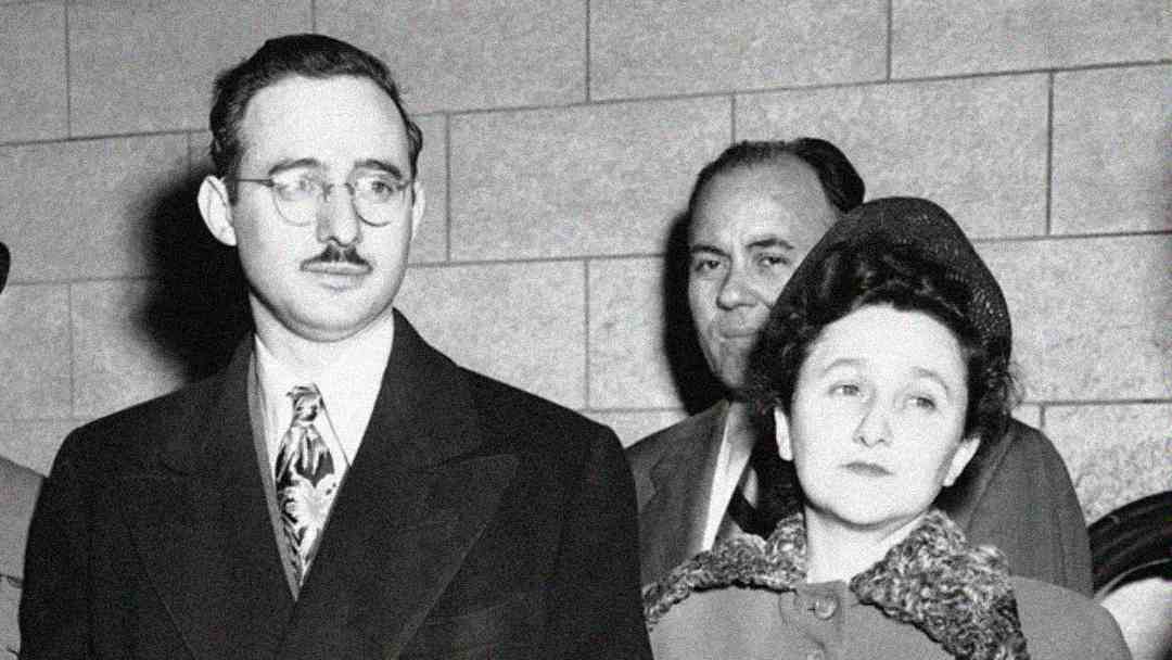 A black-and-white photo shows a man with glasses and a mustache in a suit and tie standing beside a woman in a vintage outfit with a hat, looking forward. A third man stands behind them against a tiled wall.