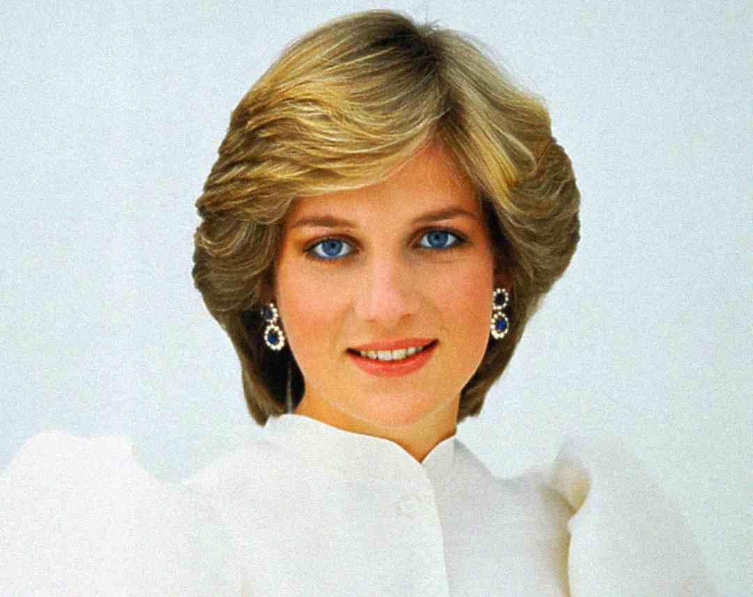 A woman with short, light brown hair and blue eyes is smiling at the camera. She is wearing a white blouse and sapphire and diamond earrings, with a light background behind her.