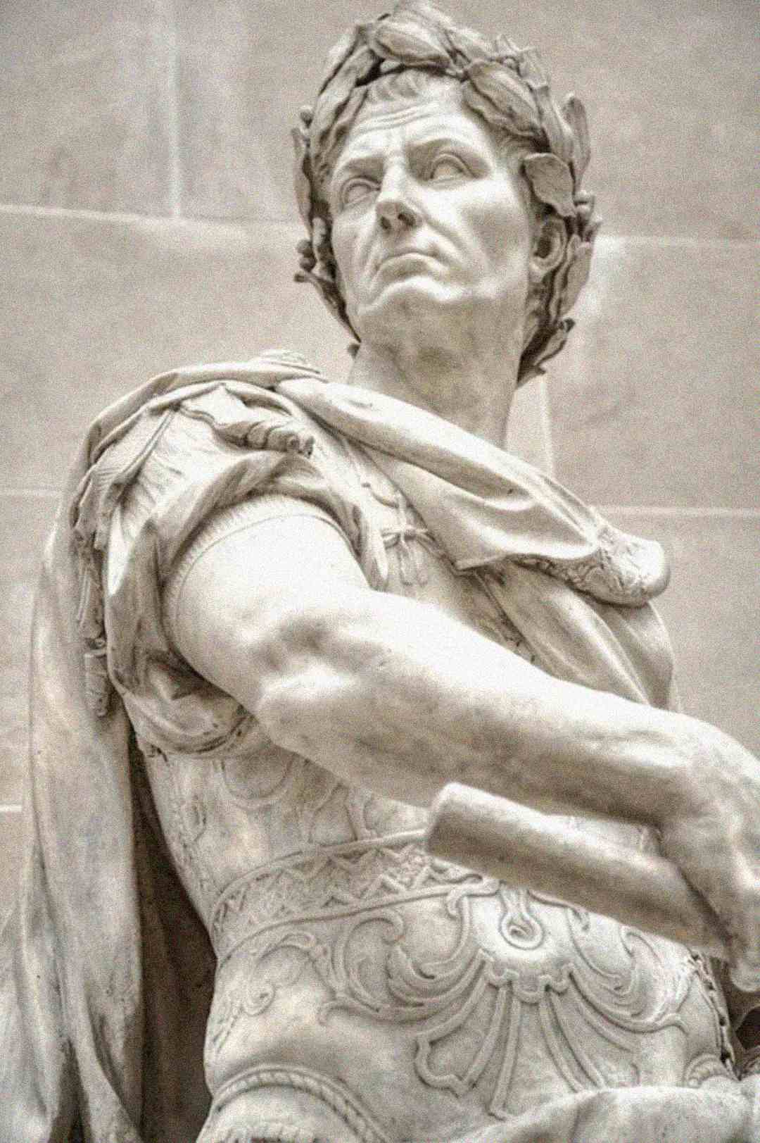 Marble statue of a man with a laurel wreath on his head, wearing ornate armor and a cloak, looking sternly to the side; classical Roman style sculpture.