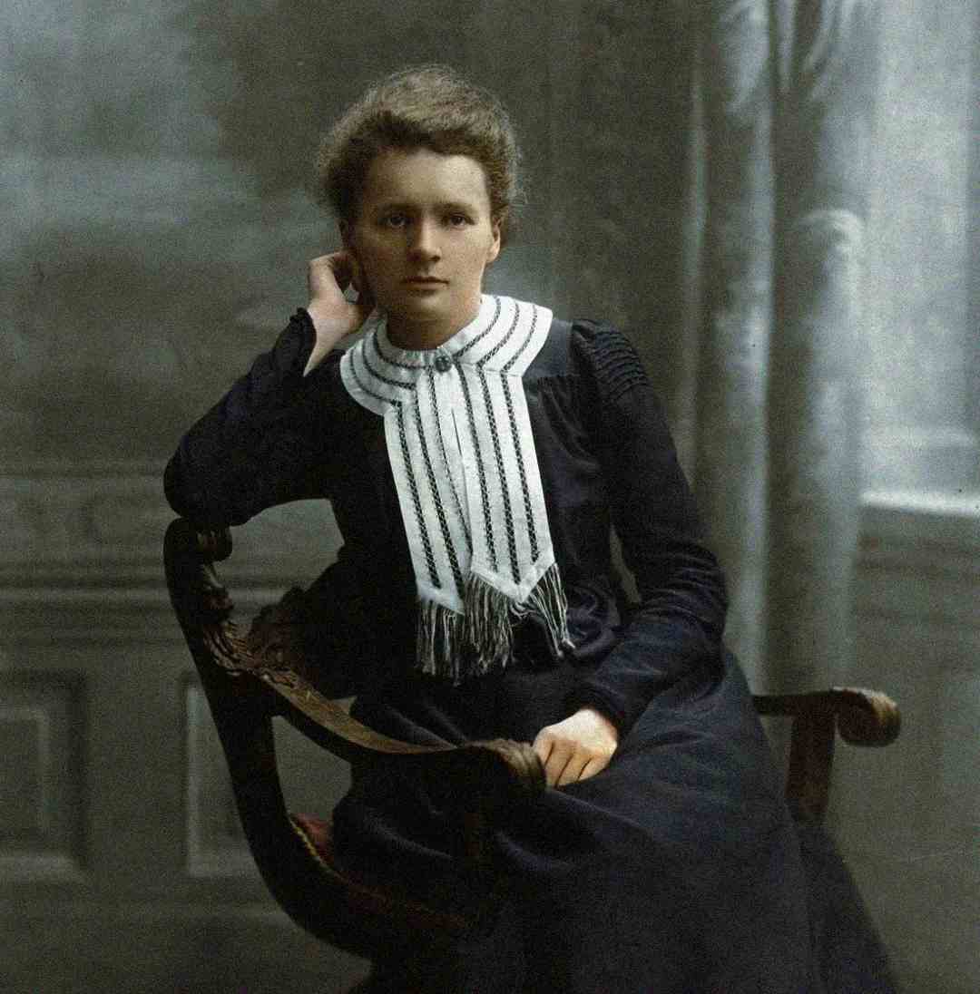 A young woman with light skin and neatly styled hair sits on an ornate chair, wearing a dark dress with a high, white, pleated collar. She rests her head on one hand and looks calmly at the camera in a vintage-style portrait.