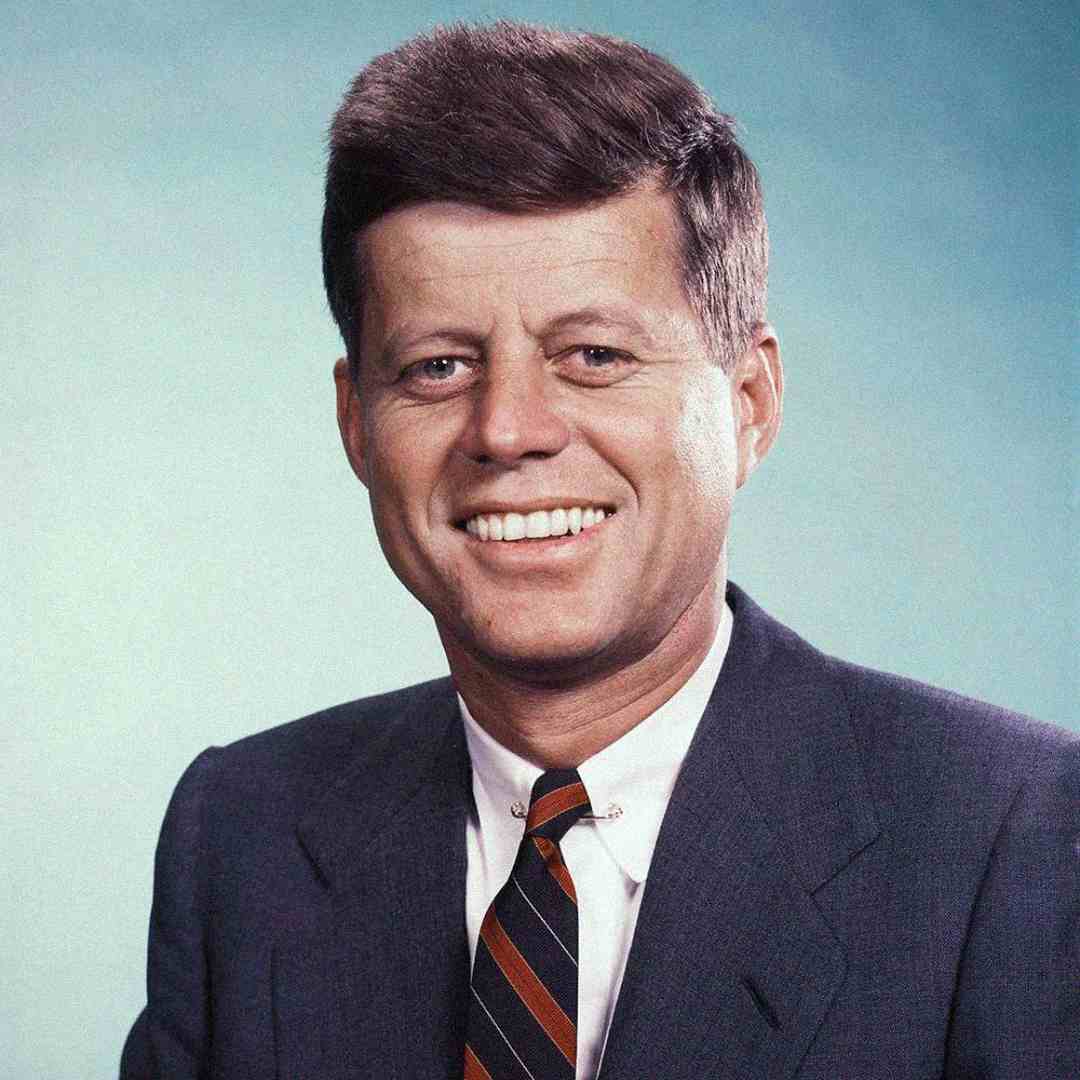 A man in a suit and striped tie smiles at the camera against a blue background. He has short, dark hair and is clean-shaven.