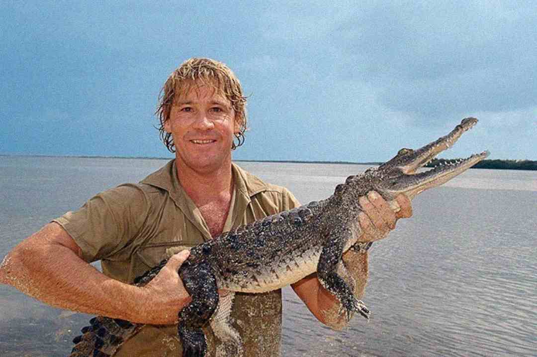 A smiling man in a khaki shirt holds a small crocodile by the water's edge, with sand on his hands and arms, and cloudy sky in the background.