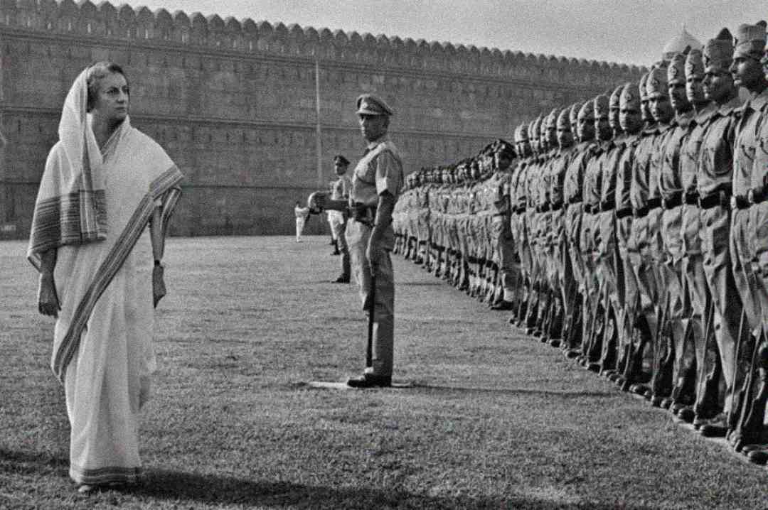 A woman in a white sari walks past a row of uniformed soldiers standing at attention on a grassy field, with a historic fort wall in the background.