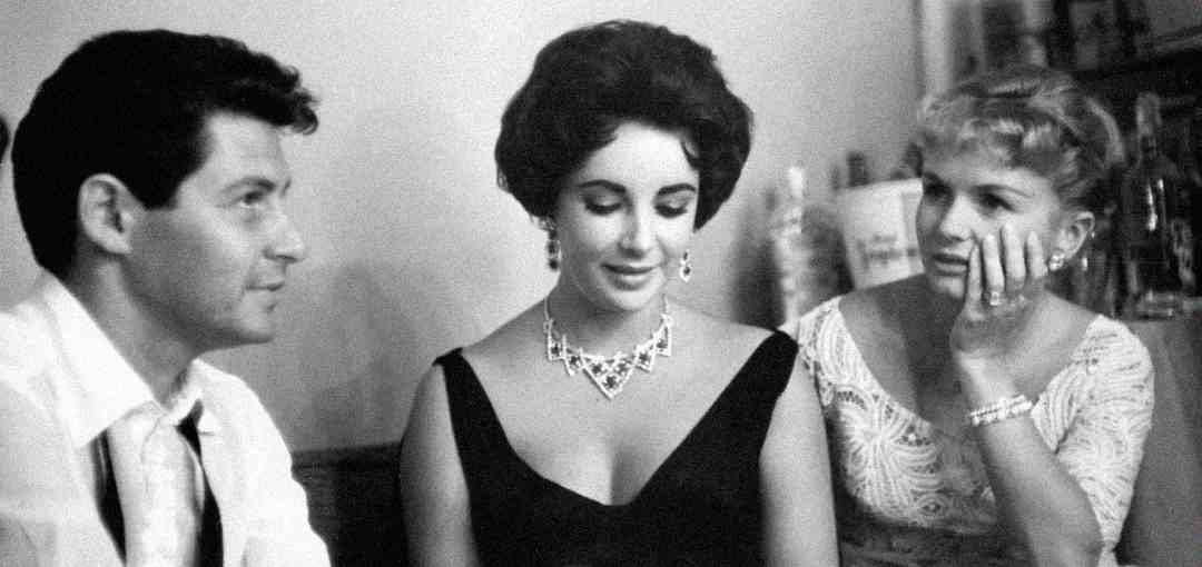 Three people sit together indoors; a man in a shirt and tie faces two women, one in a dark dress with a statement necklace, the other in a light, patterned dress, resting her head on her hand as they converse.