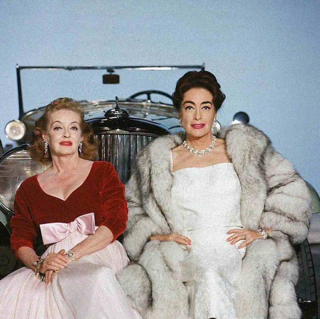 Two glamorous women sit in front of a vintage car. One wears a red velvet top with a pink skirt and bow, while the other wears a white gown and a luxurious fur coat. Both have styled hair and elegant jewelry.