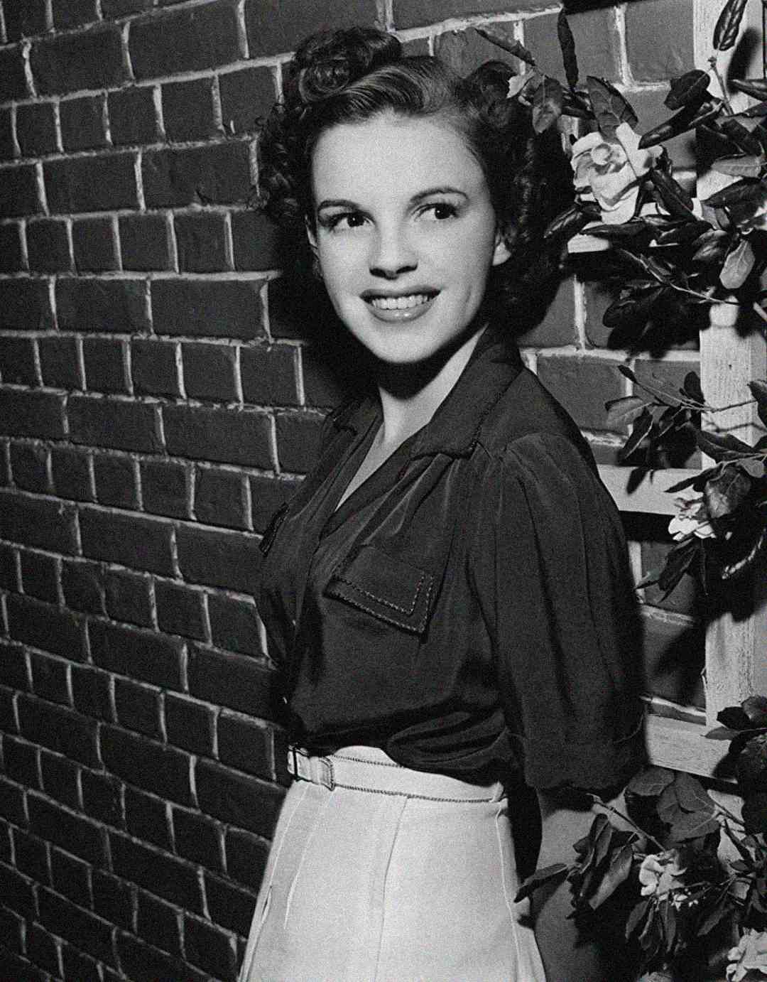 A smiling young woman with curled dark hair stands against a brick wall, wearing a dark blouse tucked into a high-waisted light skirt. Some flowers and leaves are visible on the right side of the image. The photo is in black and white.
