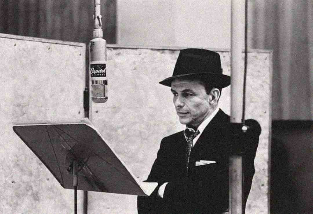 A man in a suit and hat stands at a music stand in front of an old-fashioned microphone, appearing to be in a recording studio. The image is in black and white.
