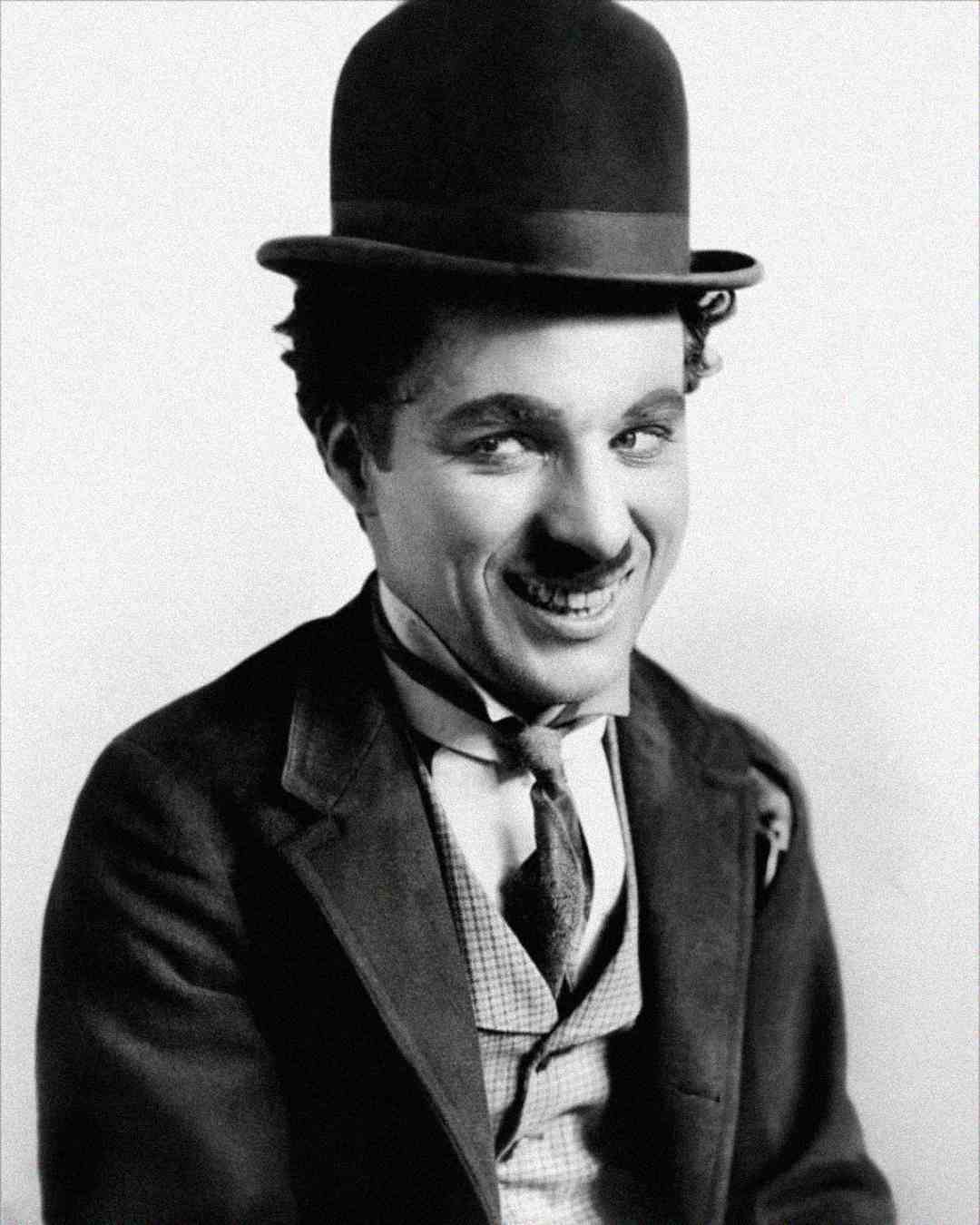 A man with a small mustache and curly hair wears a bowler hat, a suit, a vest, and a bow tie. He is smiling and looking slightly to the side against a plain background.