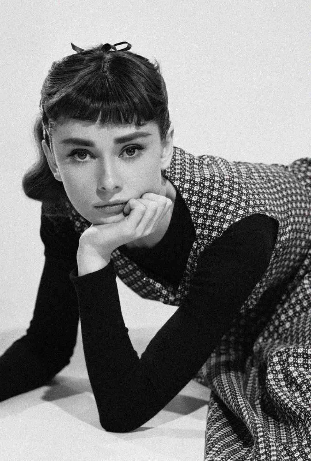 A woman with dark hair and bangs, wearing a patterned dress over a long-sleeve shirt, leans forward with her chin resting on her hand, gazing thoughtfully at the camera in a black-and-white photo.