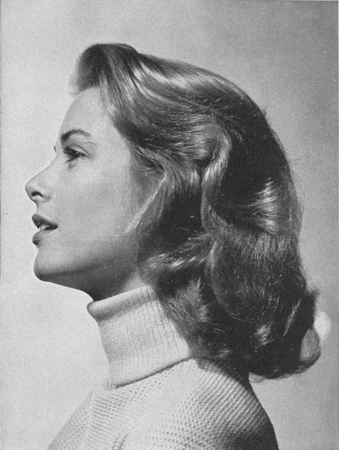 Black and white portrait of a woman in profile with wavy, shoulder-length hair, wearing a light-colored turtleneck sweater and looking left against a plain background.