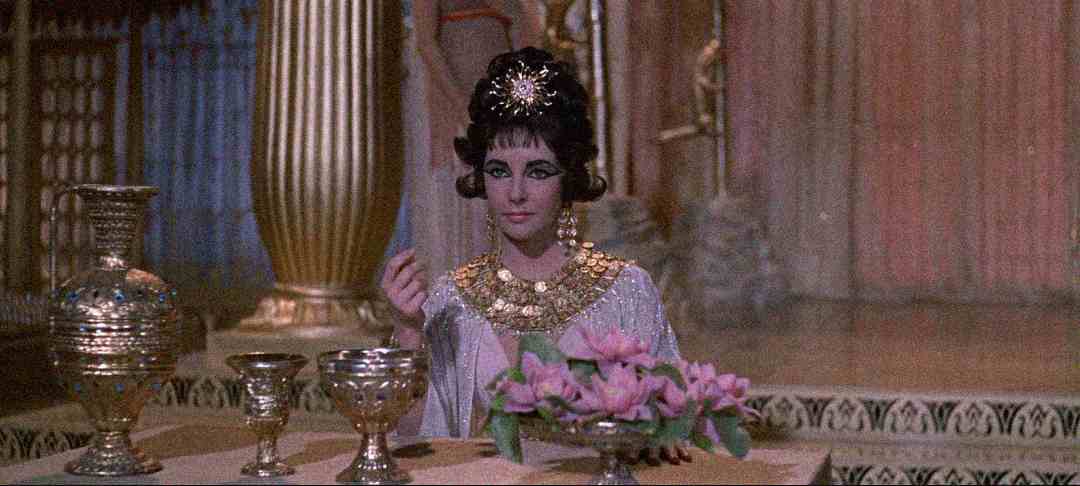 A woman dressed as Cleopatra sits at an ornate table with golden goblets and a vase of pink flowers, wearing an elaborate gold necklace and headpiece, in a richly decorated ancient Egyptian-style room.