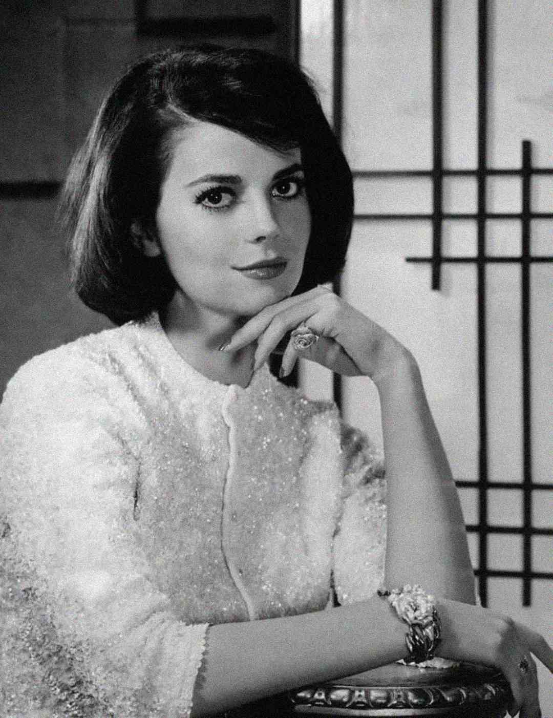 A woman with dark, shoulder-length hair and a soft expression poses in a sparkly, light-colored top, resting her chin on her hand. Geometric lines decorate the background in this black-and-white portrait.