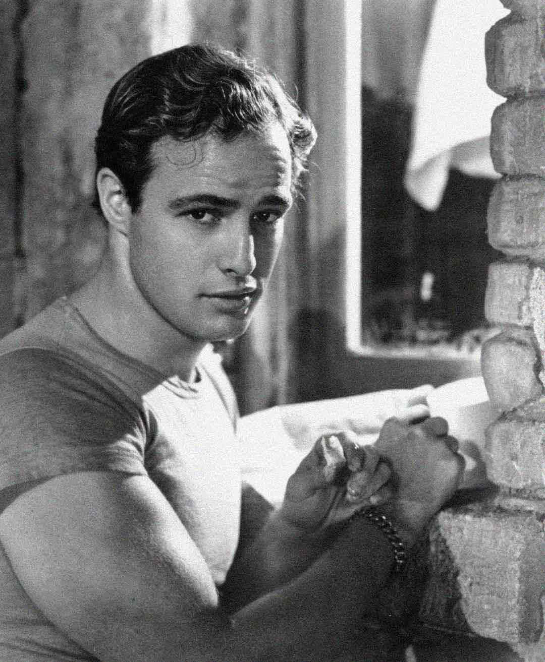 A young man with wavy hair and a fitted t-shirt stands by a brick wall, looking directly at the camera with a serious expression, hands clasped together. The photo is black and white.