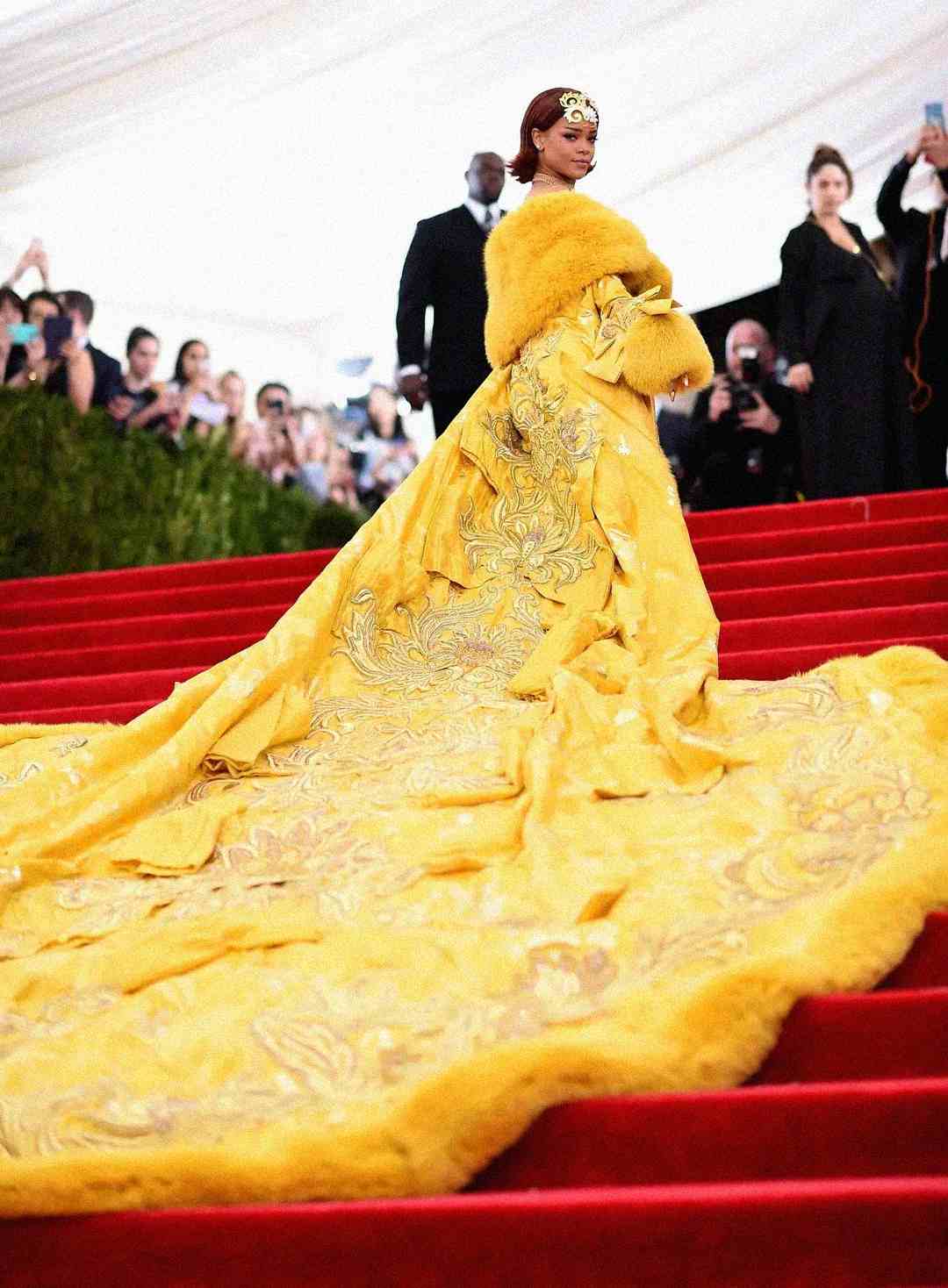 A woman in a lavish yellow gown with a long embroidered train stands on red carpeted stairs, surrounded by onlookers and photographers at a formal event.