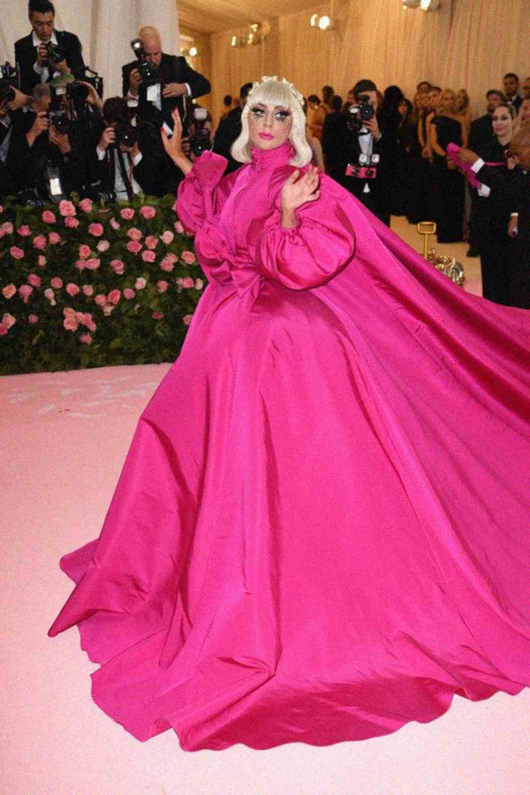 A person poses on a pink carpet wearing a dramatic, voluminous, bright pink gown with puffy sleeves. Photographers and flowers are visible in the background.