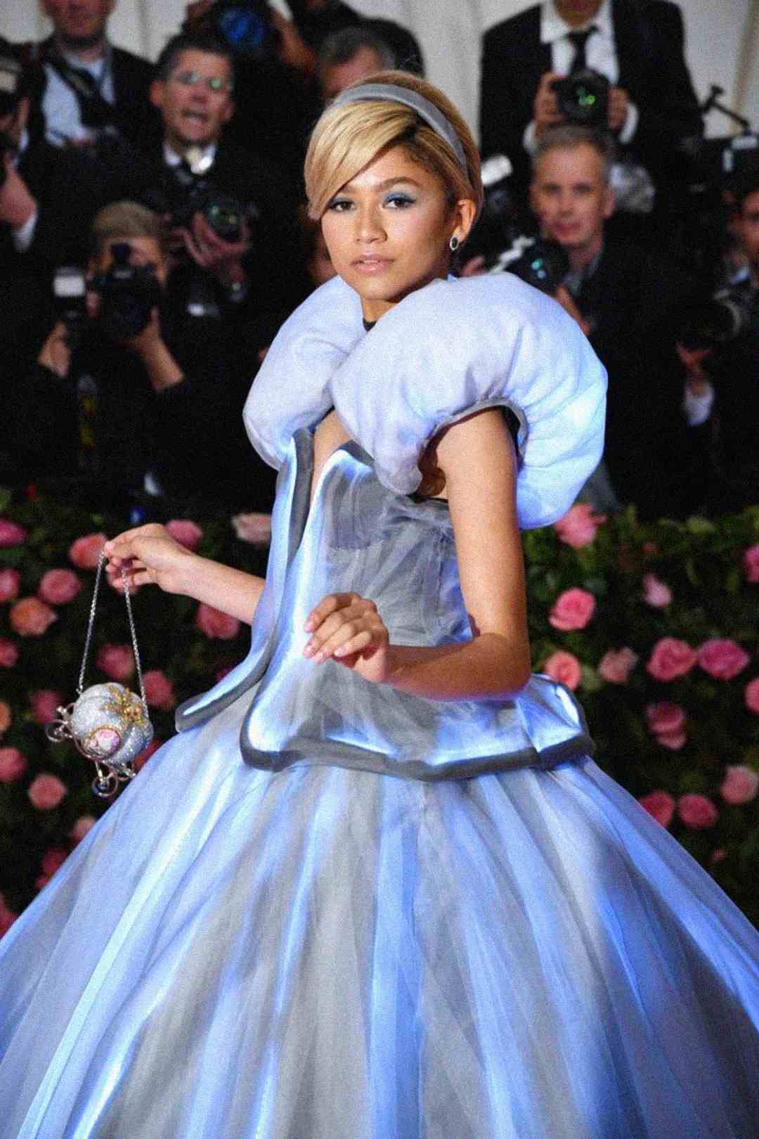 A woman in a glowing, futuristic blue ball gown with a high, rounded collar walks the red carpet, surrounded by photographers and pink rose decorations in the background. She holds a small, illuminated bag.