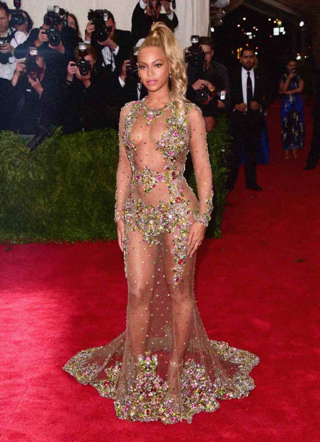 A woman poses on a red carpet in a sheer, embellished gown adorned with sparkling floral details. Photographers stand in the background capturing her photo.