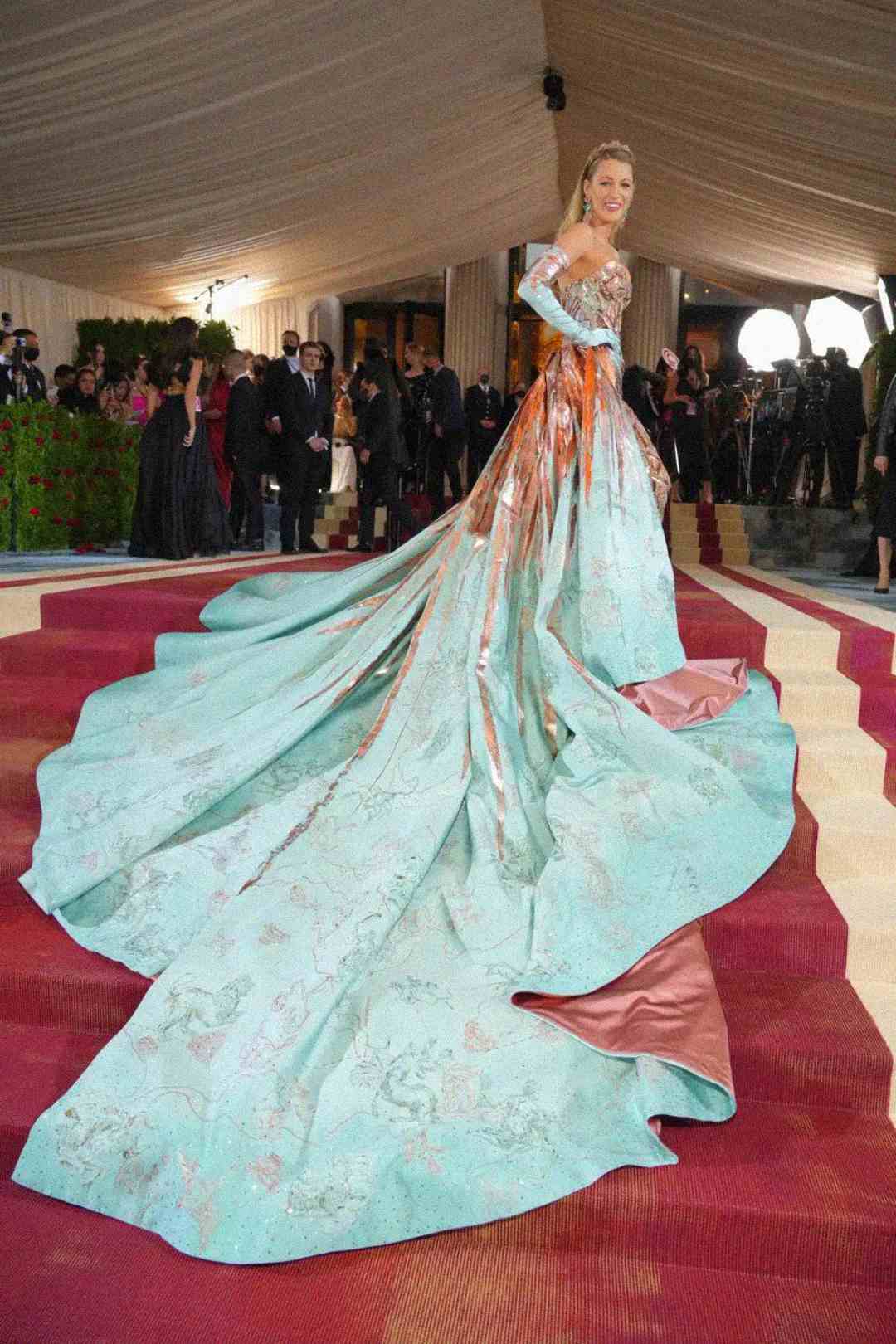 A woman poses on a red carpet wearing a dramatic aqua-blue gown with metallic patterns and an elaborate train, surrounded by photographers and guests in a tented venue.