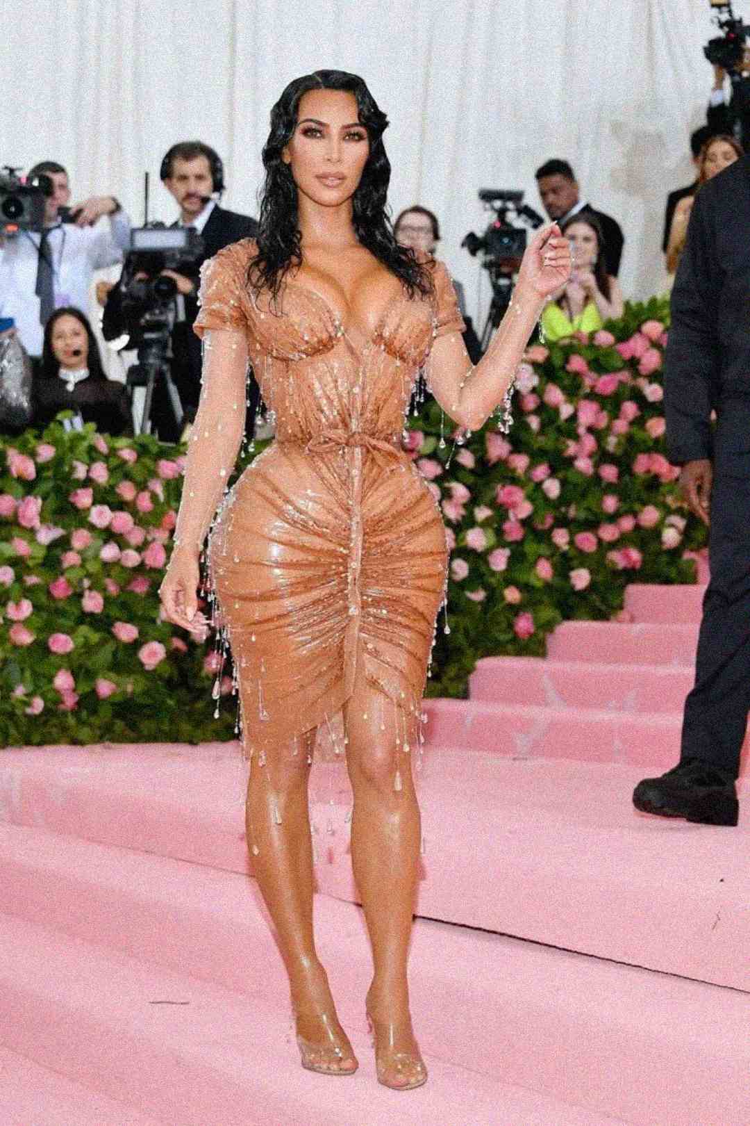 A woman in a nude-colored, form-fitting, wet-look dress adorned with crystal droplets stands on pink carpeted stairs, surrounded by photographers and pink rose decorations at a glamorous event.