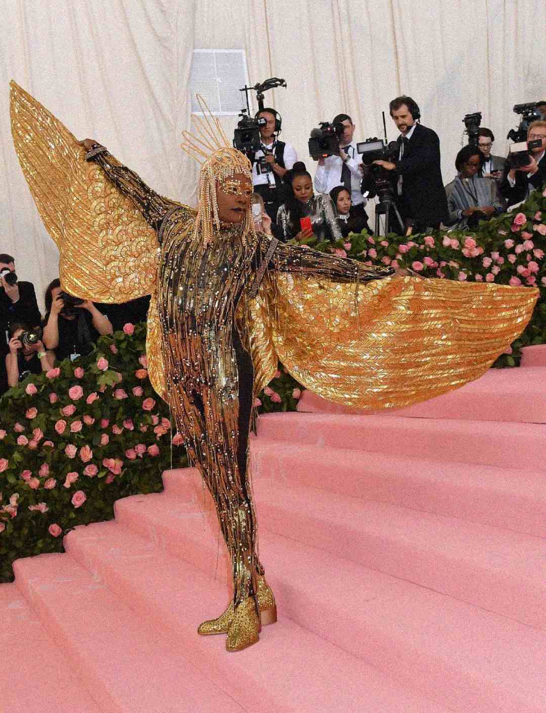 A person poses on pink stairs in a dramatic gold outfit with large wing-like sleeves, gold boots, and a gold headpiece. Photographers and onlookers stand in the background, surrounded by pink roses.