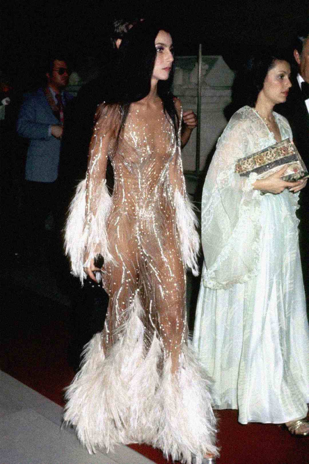 A woman in a sheer, nude-colored gown adorned with sparkling beads and white feather cuffs and hem walks on a red carpet, accompanied by others in formal evening attire.