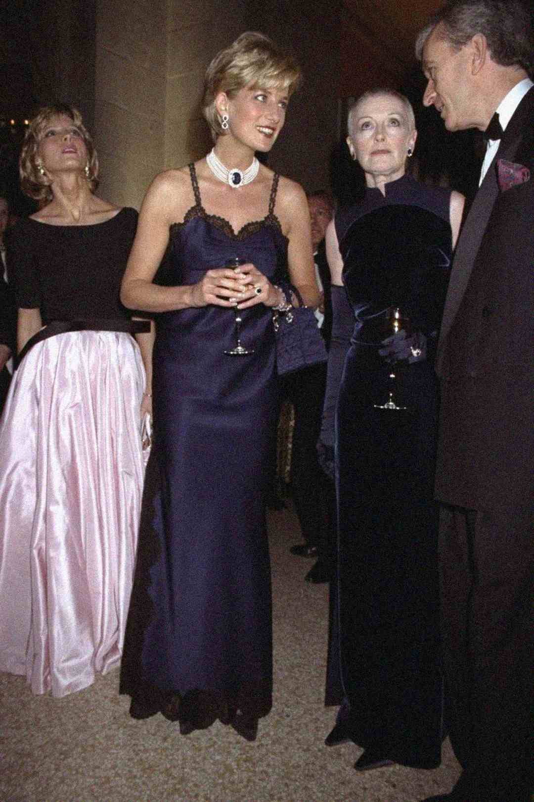 Four elegantly dressed people at a formal event, with two women in dark evening gowns holding wine glasses and conversing with a man in a tuxedo, while another woman in a pink skirt glances upward.